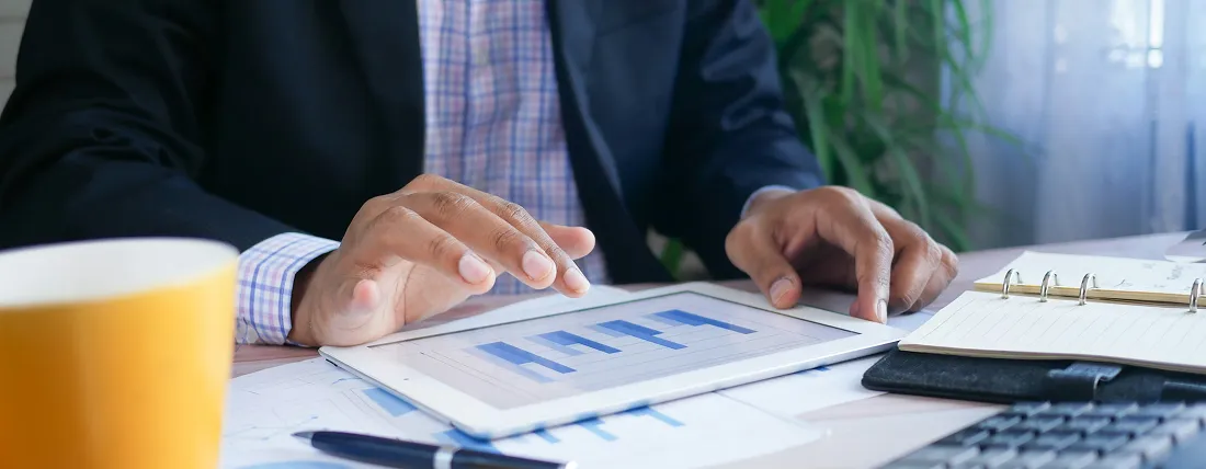 Person in business attire analyzing bar charts on a tablet at a desk with papers and a notebook.