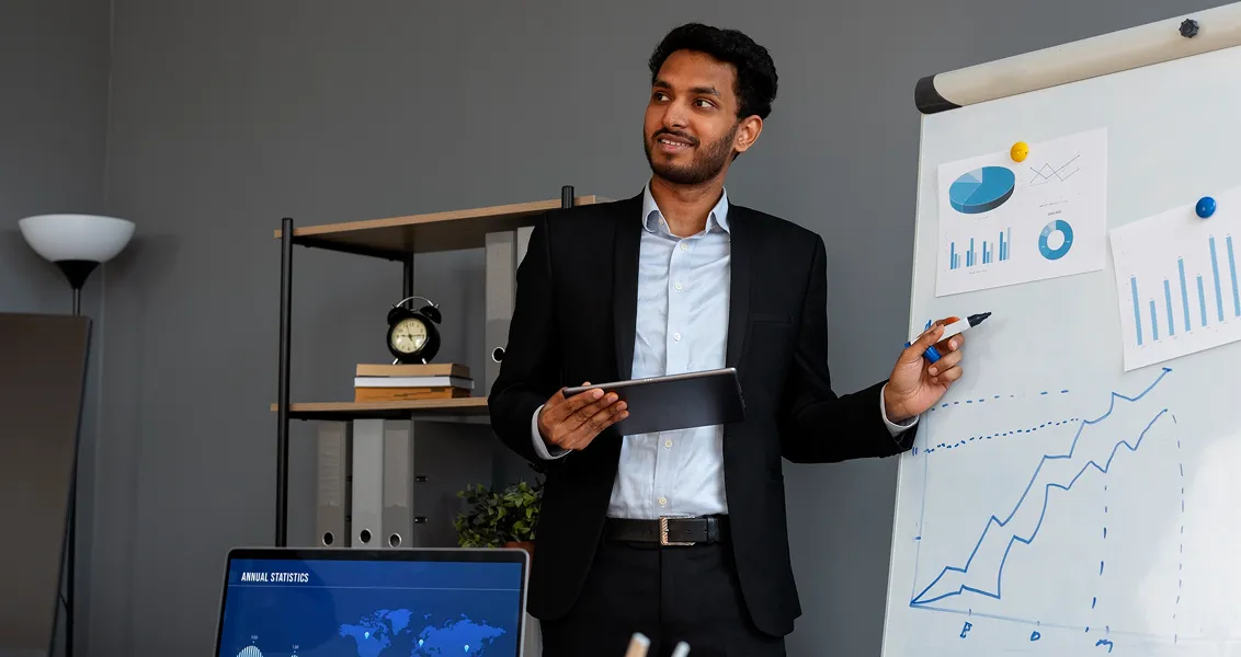 Man in black suit holding tablet, pointing at chart with rising graphs on whiteboard in office.