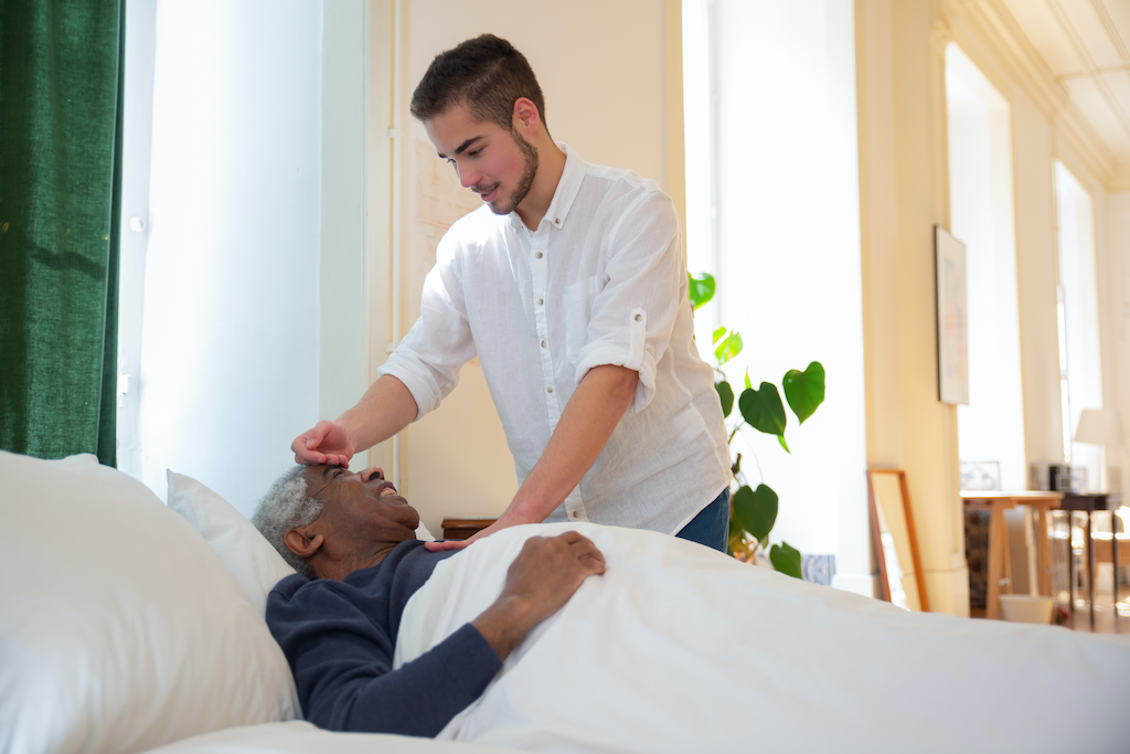 a masculine presenting person checking the temperature of a racialized person of advanced age laying on bed