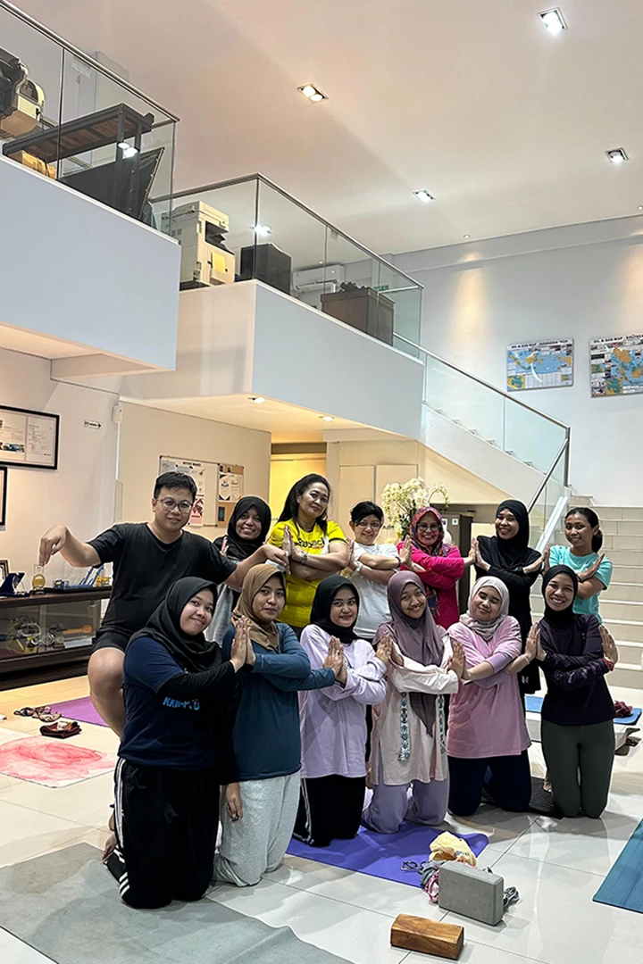 Group of eleven women and one man posing indoors on yoga mats with palms pressed together, smiling at the camera.