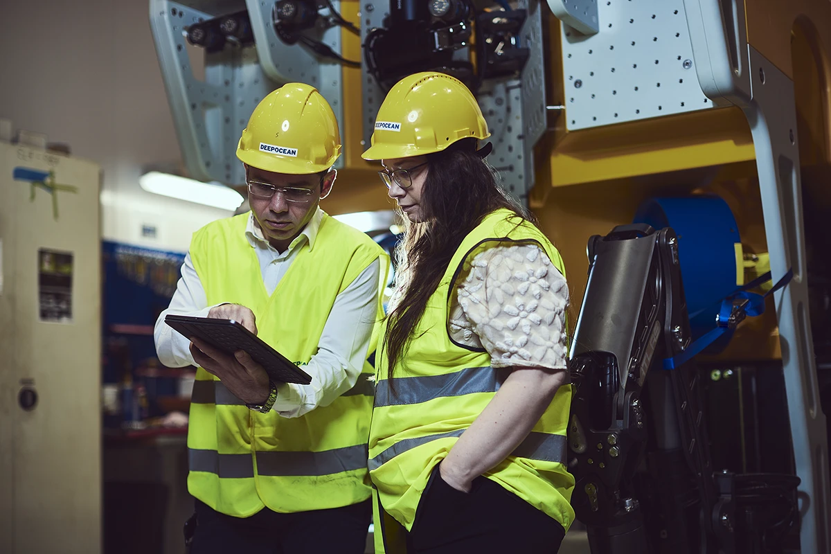 Two workers wearing yellow hard hats and high-visibility vests reviewing information on a tablet in an industrial setting.