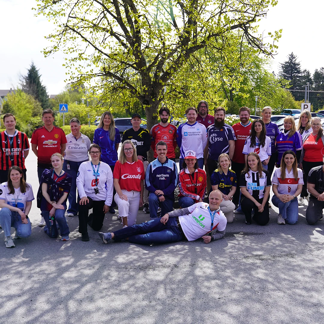 Group of 23 people wearing various football jerseys posing outdoors under a leafy tree on a sunny day.