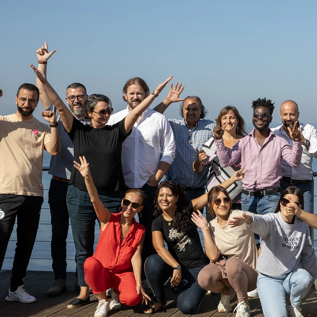 Group of diverse adults outdoors on a sunny day, smiling and posing energetically on a wooden deck by the water.