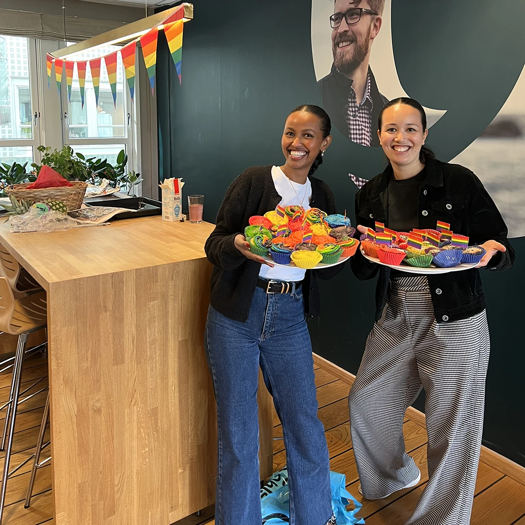 Two smiling women holding plates of colorful rainbow cupcakes with small pride flags, standing in a decorated room with rainbow bunting.
