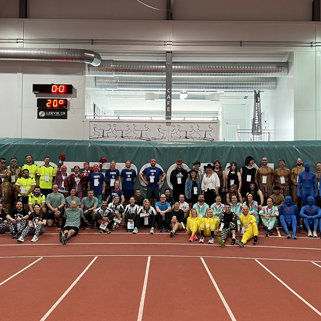 Large group of people in various colorful costumes and uniforms posing together inside a sports track facility with a digital scoreboard showing 0:0 and 20°C.