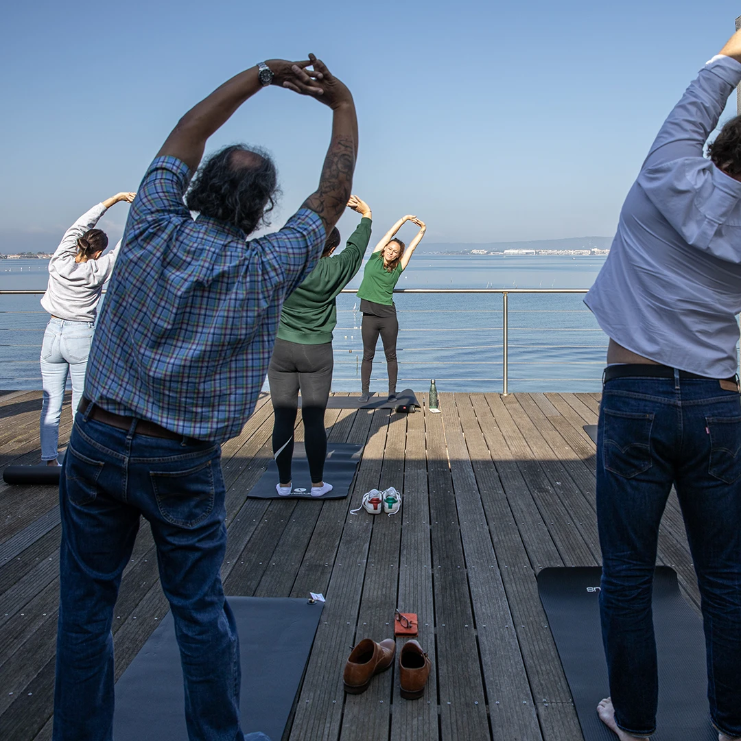 Group of people stretching on yoga mats on a wooden deck overlooking a calm body of water under clear blue sky.