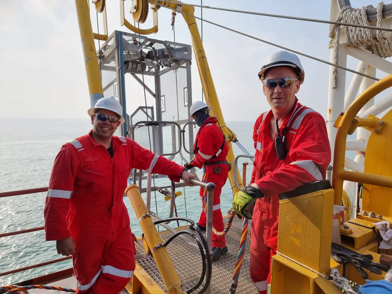 Three offshore workers in red safety suits and helmets on a platform above the sea.
