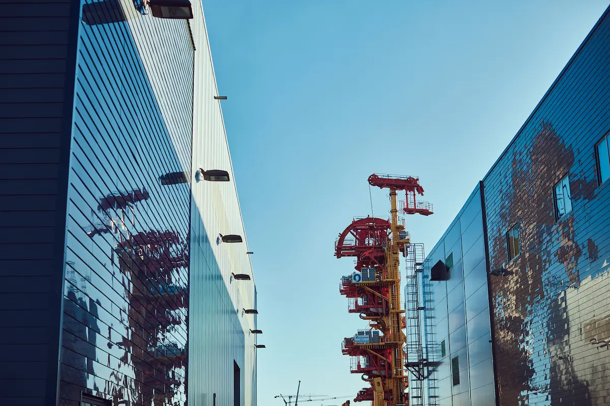 Industrial red and yellow metal structure between two reflective warehouse buildings under blue sky.