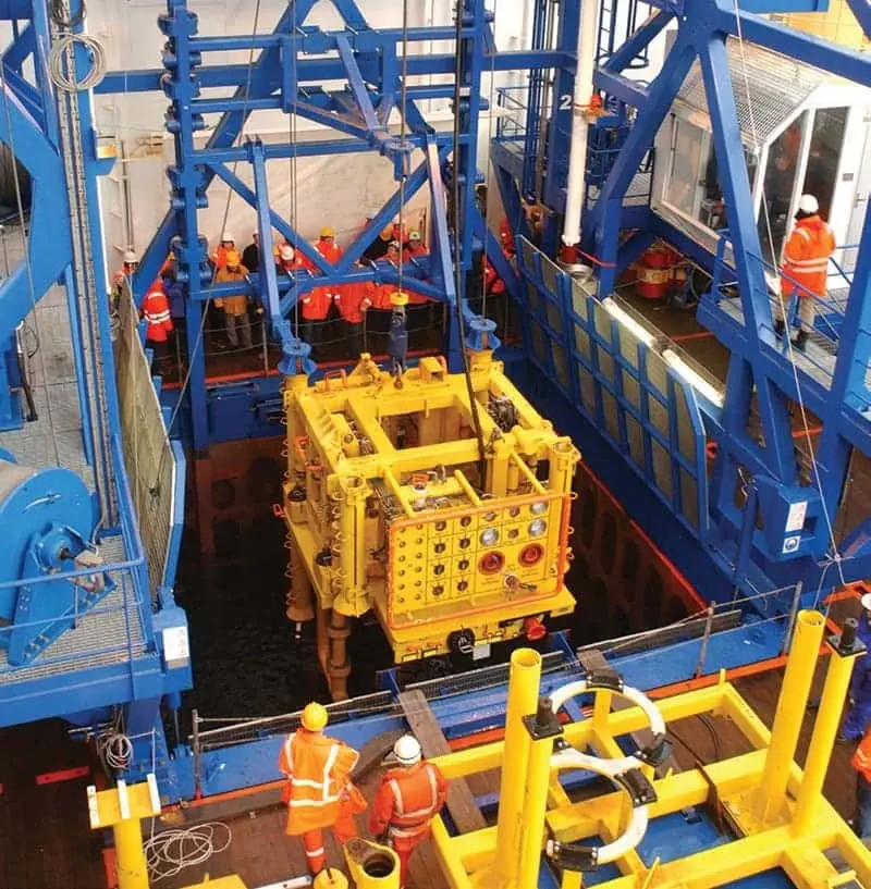 Workers in orange safety gear operate blue and yellow offshore equipment on an oil rig platform.