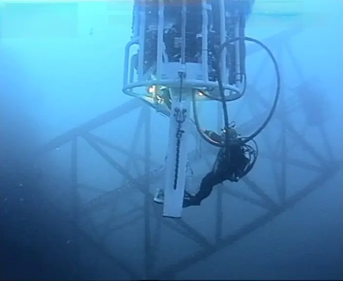 Scuba diver inspecting or working on an underwater metal structure with equipment suspended above.