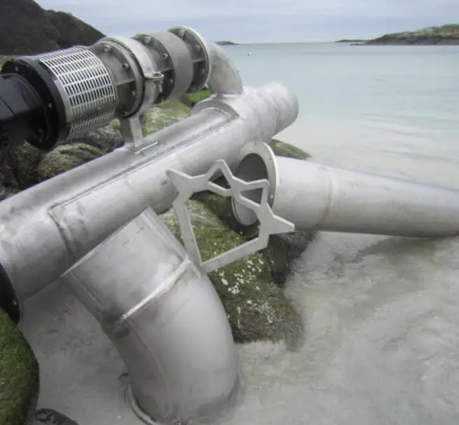 Metal pipes with a valve on a rocky shore extending into the water under a cloudy sky.