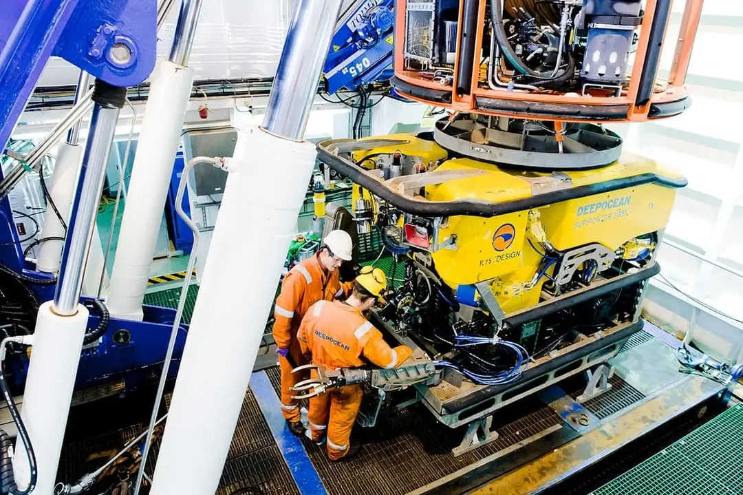 Two workers in orange coveralls inspecting a yellow underwater vehicle labeled DeepOcean indoors.