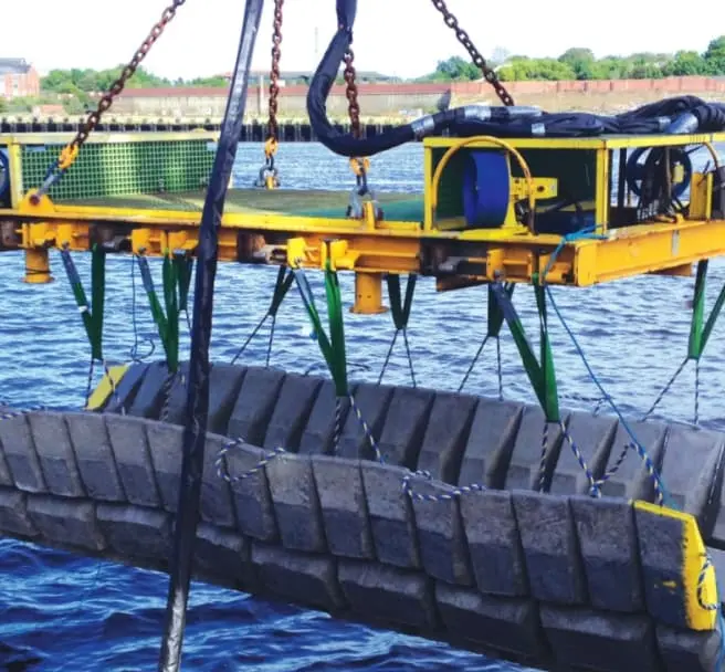 Yellow crane lifting a large segmented concrete block over water with a shoreline in the background.