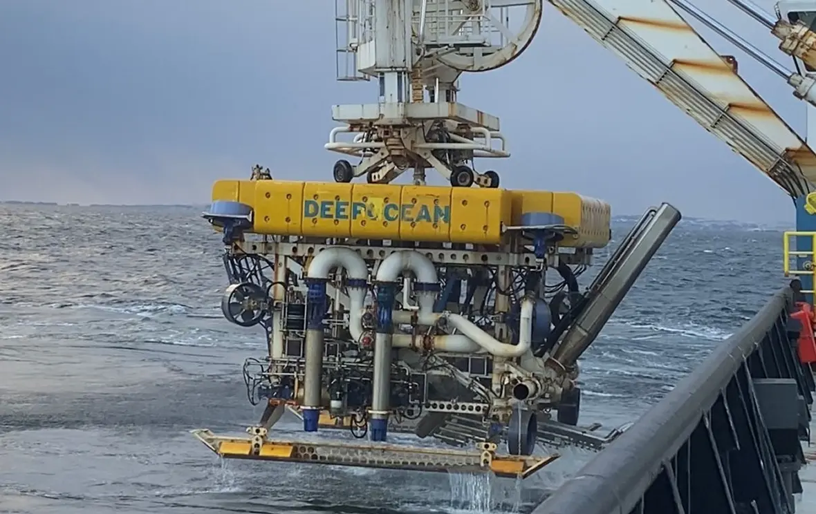 Yellow deep ocean submersible being lowered from a ship with a helicopter flying nearby above the sea.