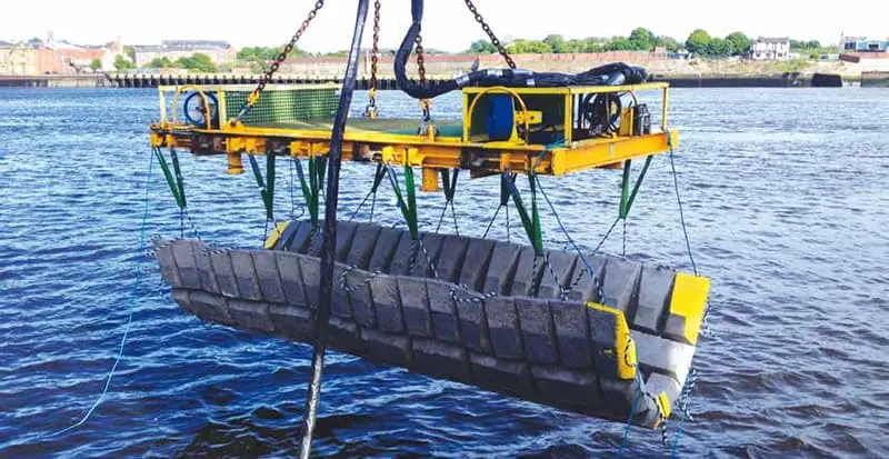 Yellow mechanical device lifting a concrete structure above water with land and buildings in the background.