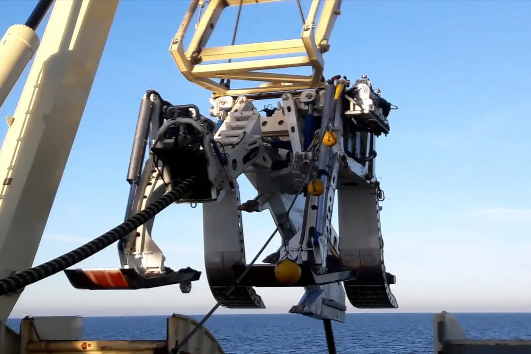 Large mechanical underwater device with cables, suspended above water from a crane on a ship.