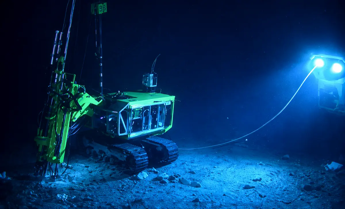 Underwater remotely operated vehicle with yellow frame working on seabed under bright blue lights.