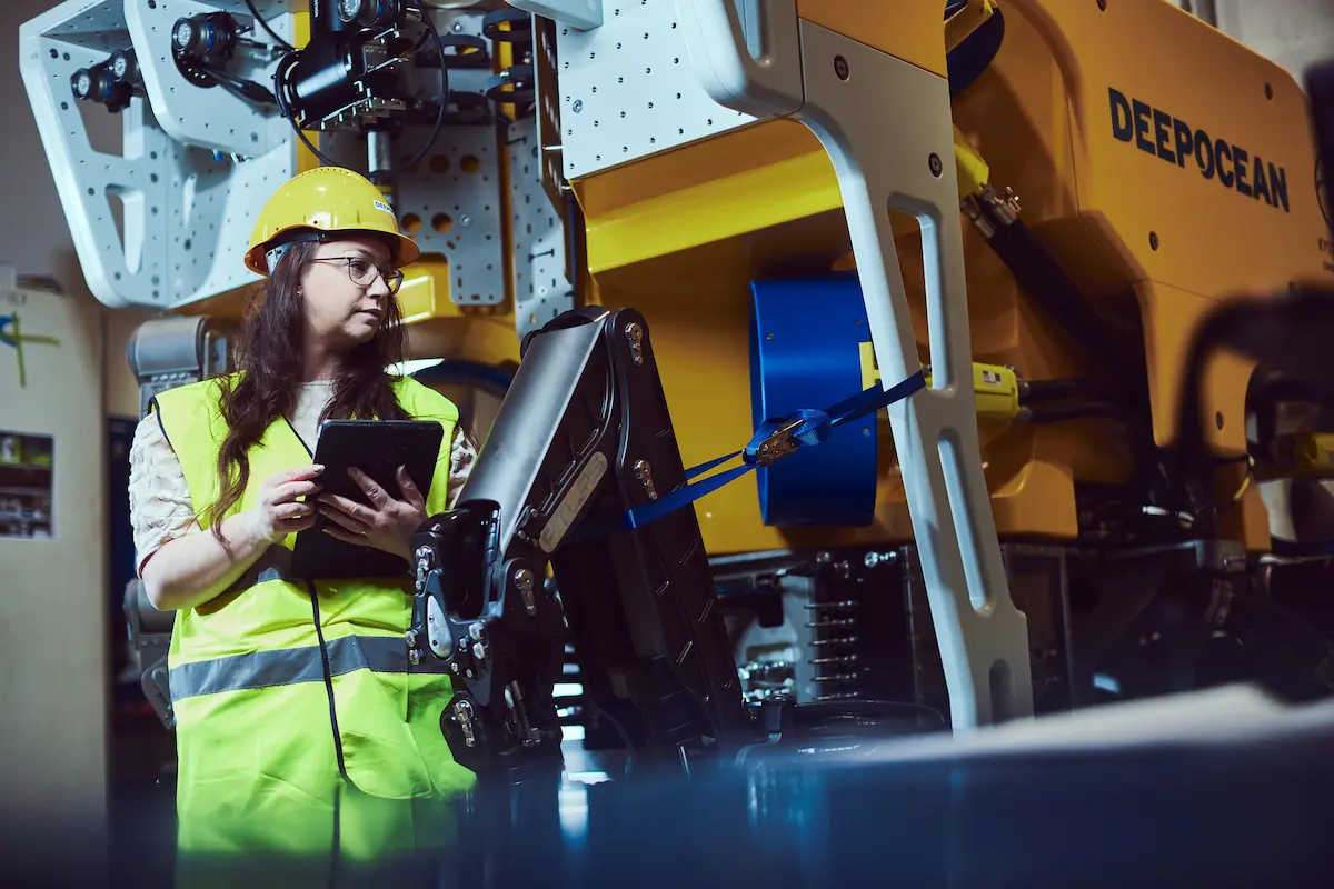 Woman in a yellow hard hat and vest using a tablet near large yellow DEEPOCEAN machinery.