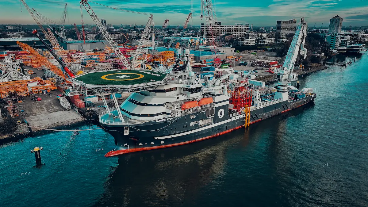 Large offshore vessel Olympic Aries docked at harbor with cranes and industrial buildings nearby.