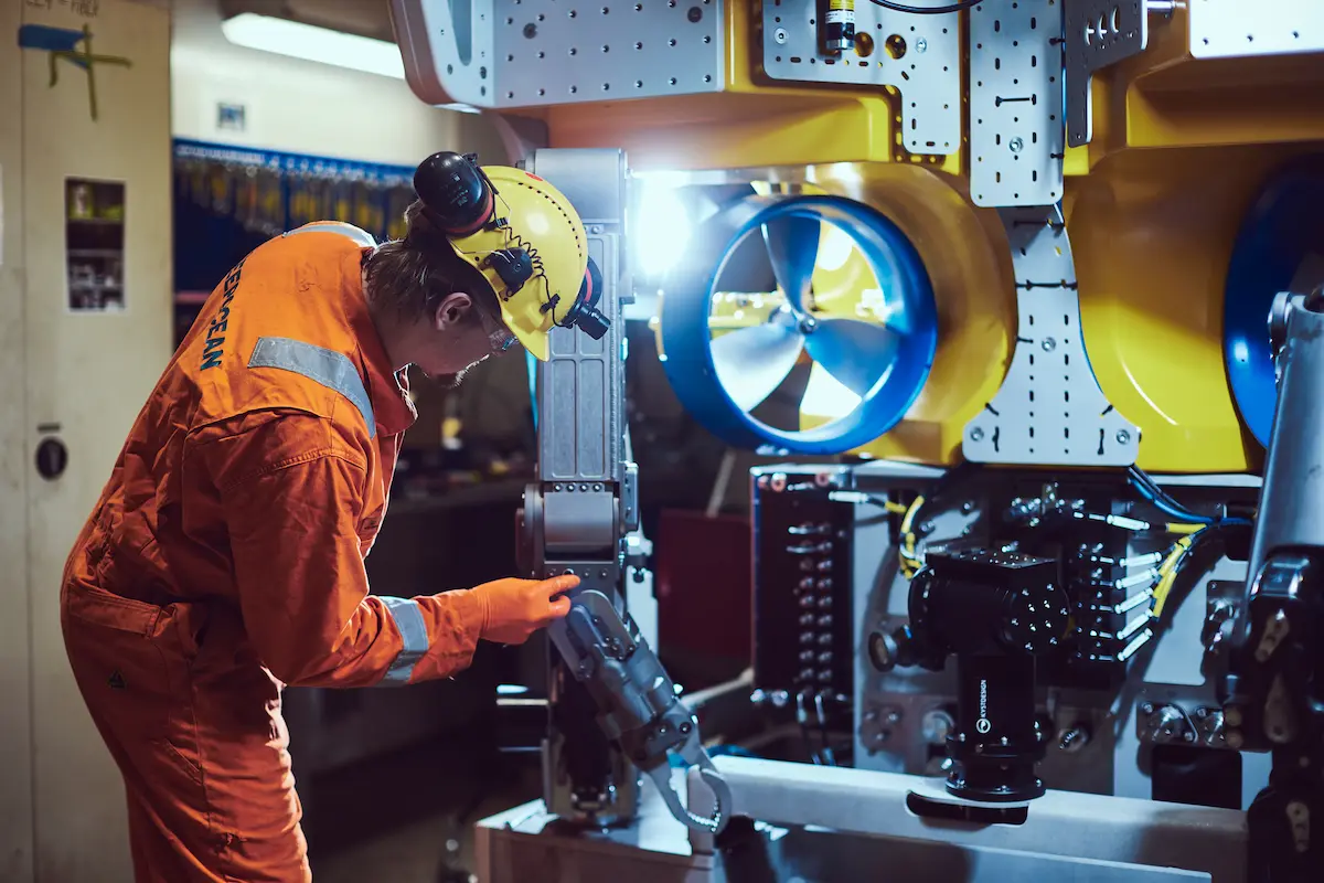 Worker in orange coverall and helmet inspecting underwater robotic equipment with large blue propellers.