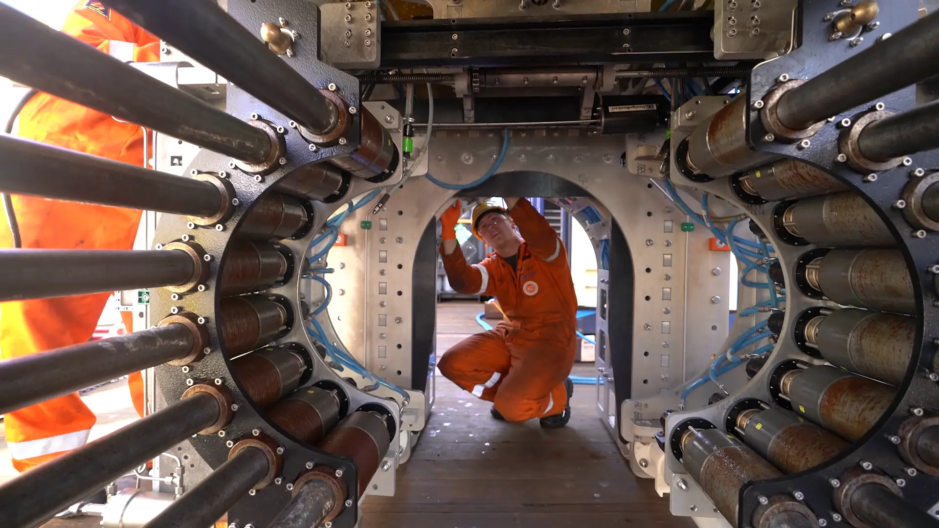 Worker in orange coveralls and helmet kneeling and inspecting a large industrial machine part.