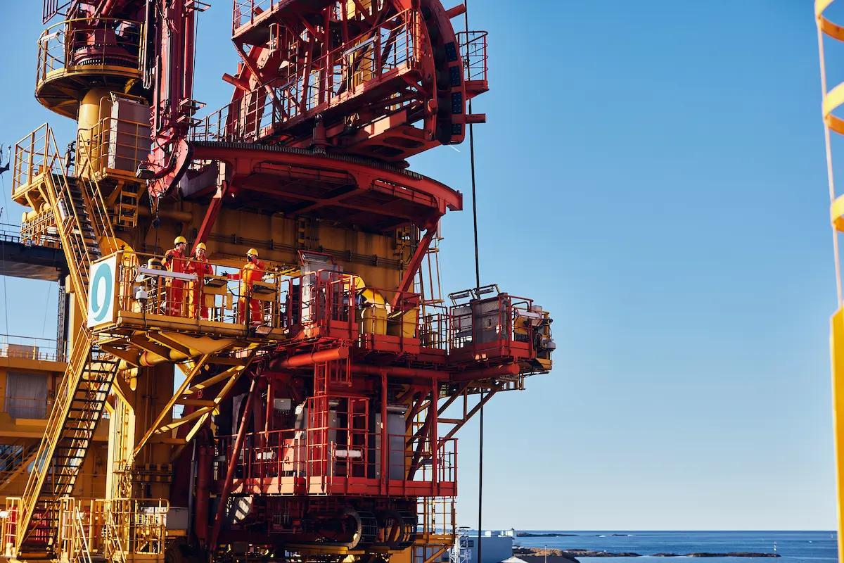 Workers in safety gear on red and yellow offshore oil rig platform with ocean in the background.