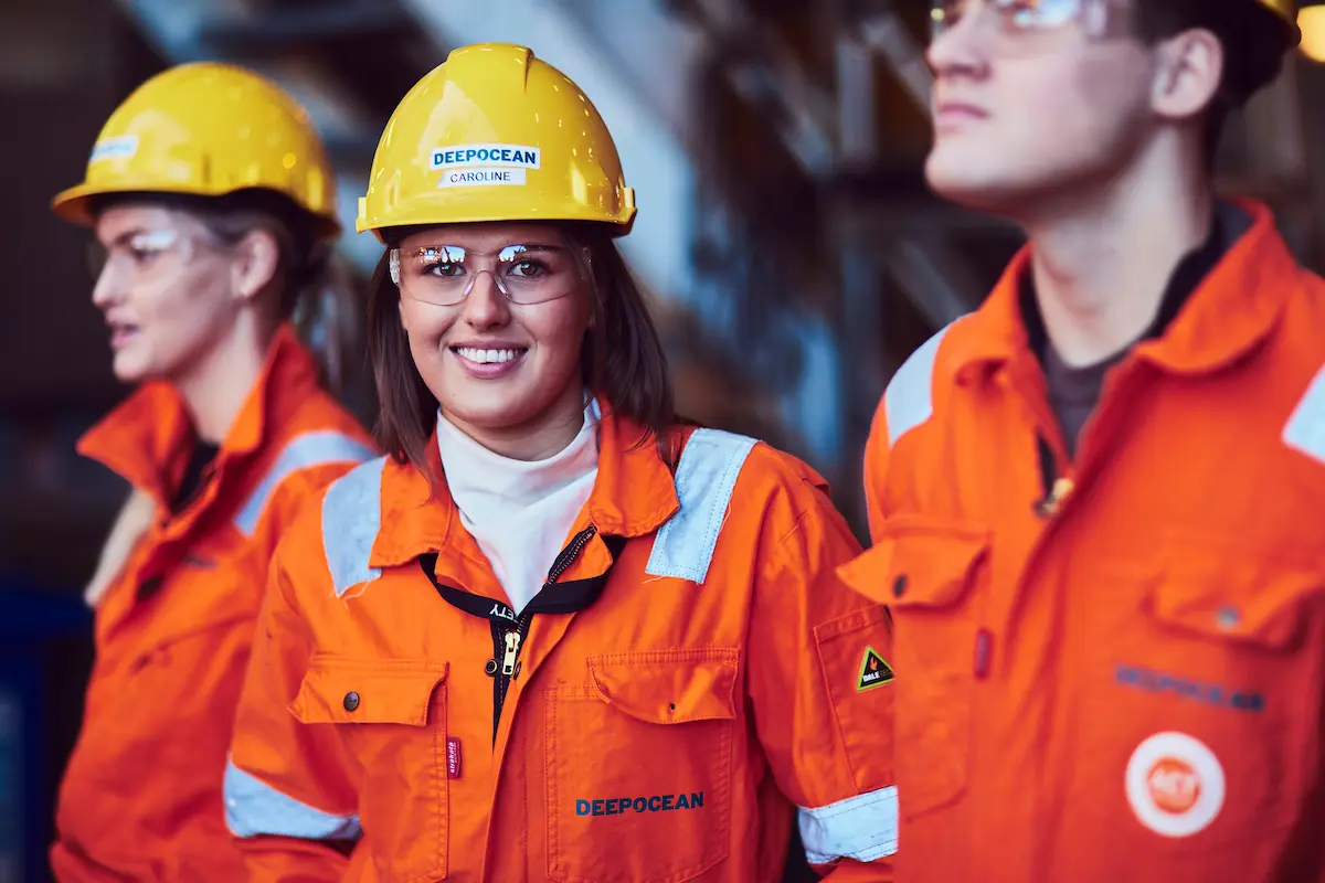 Three workers in orange safety uniforms and yellow helmets, one smiling at the camera.