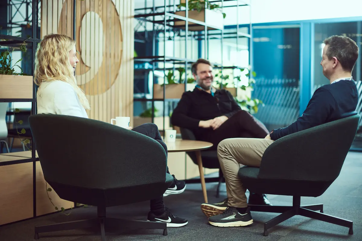 Three people sitting in black chairs around a table in a modern office with plants and wooden decor.