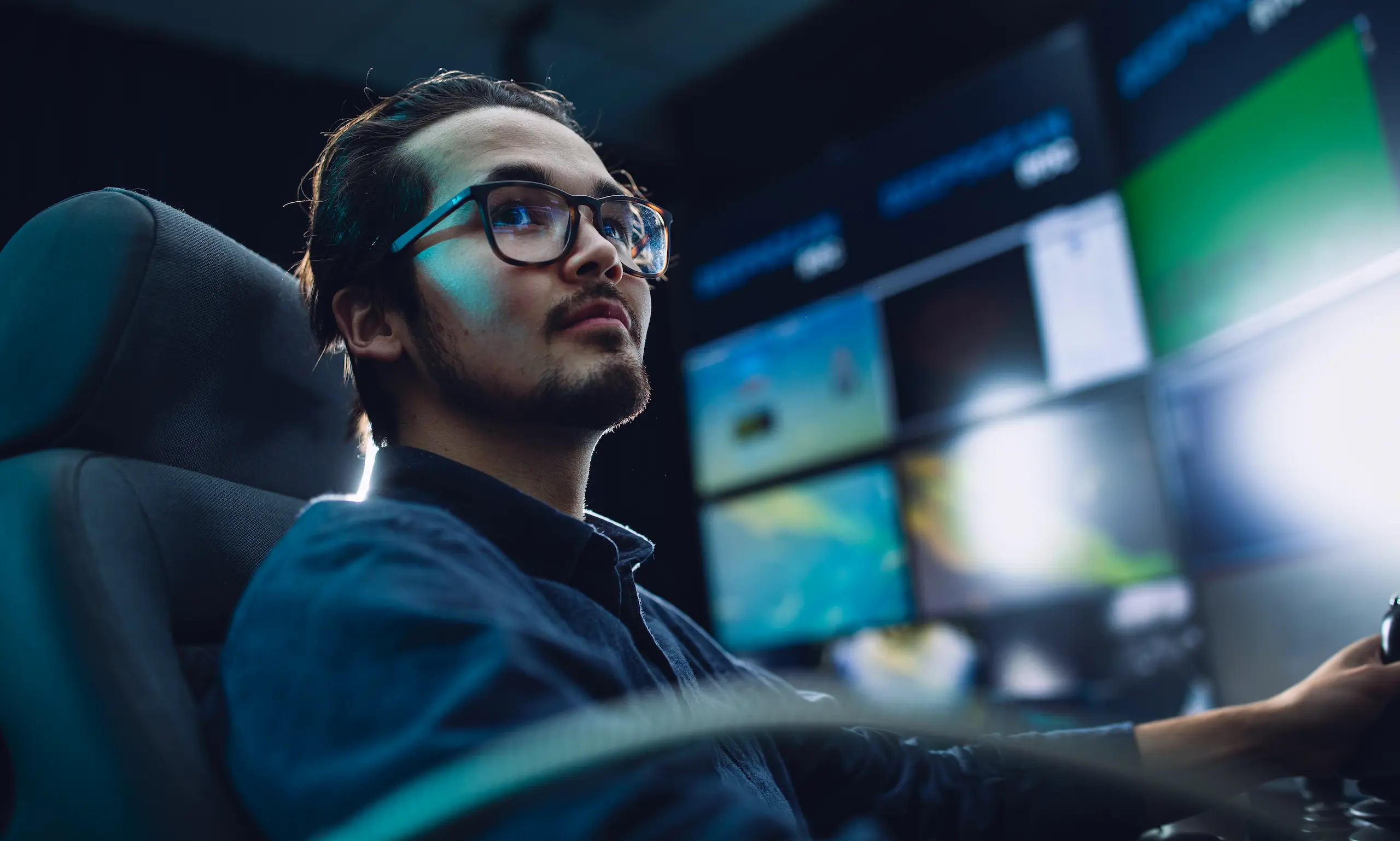 Man with glasses focused on screens in a dark room, sitting in a chair with control devices.