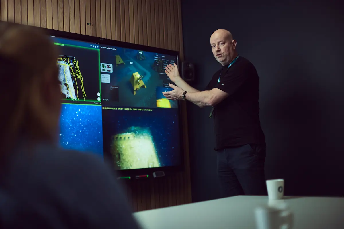 Man in black shirt presenting underwater exploration images on a large screen to a seated woman.