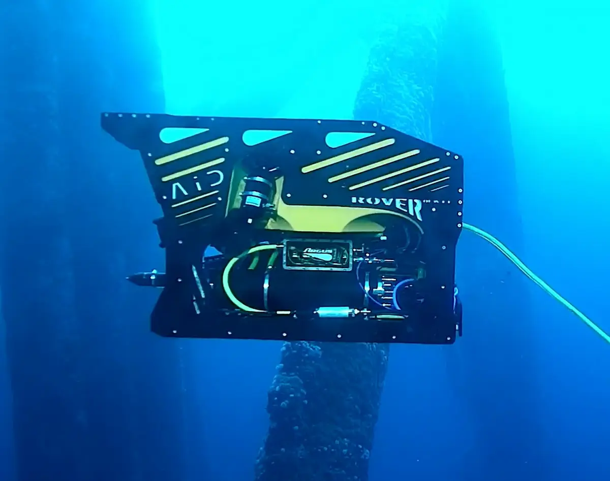 Black and yellow underwater drone inspecting marine structure covered in barnacles.