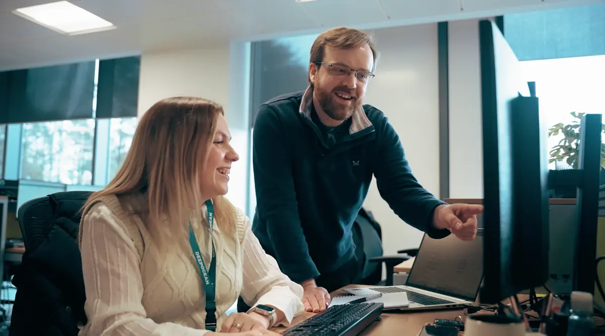 Man points at computer screen as woman smiles, both engaged in a discussion at an office desk.