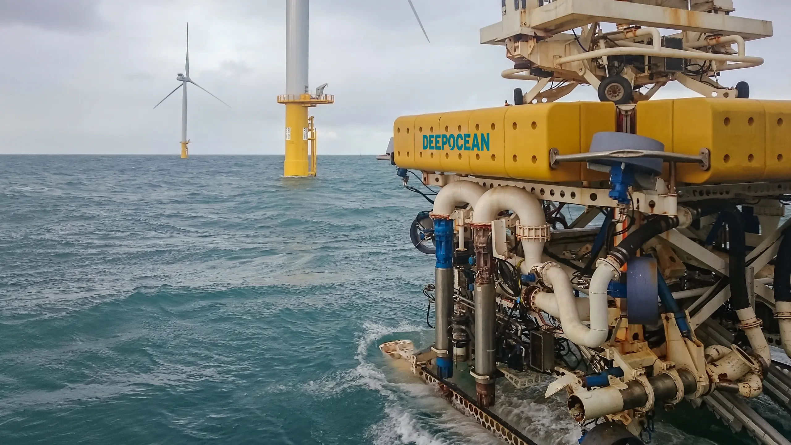 Yellow subsea intervention vehicle labeled DEEP OCEAN partially submerged near offshore wind turbine foundations in a grey, cloudy sea.