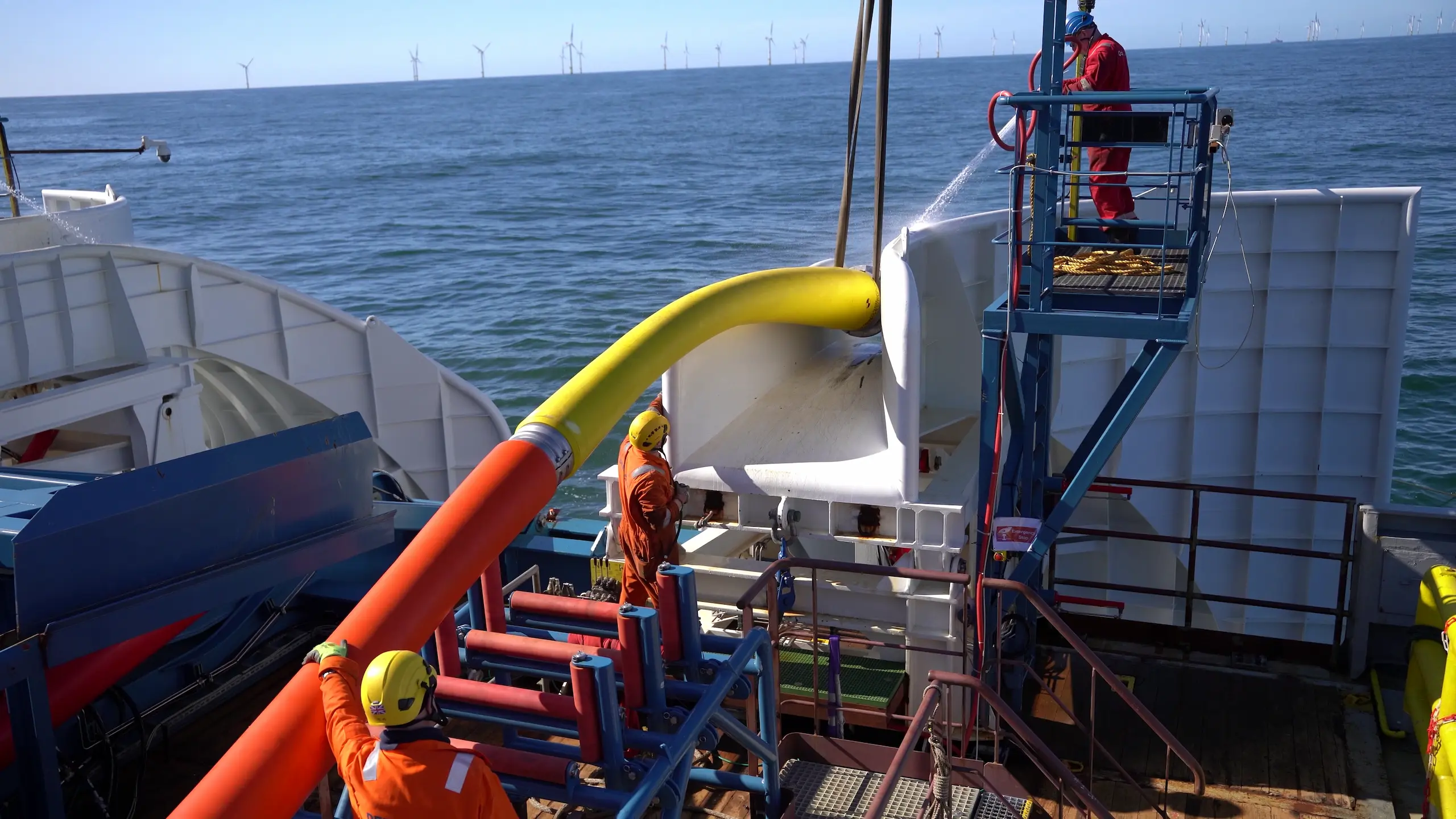 Two workers in orange safety suits and yellow helmets managing thick orange and yellow cables on a ship deck with a blue sea and offshore wind turbines in the background.