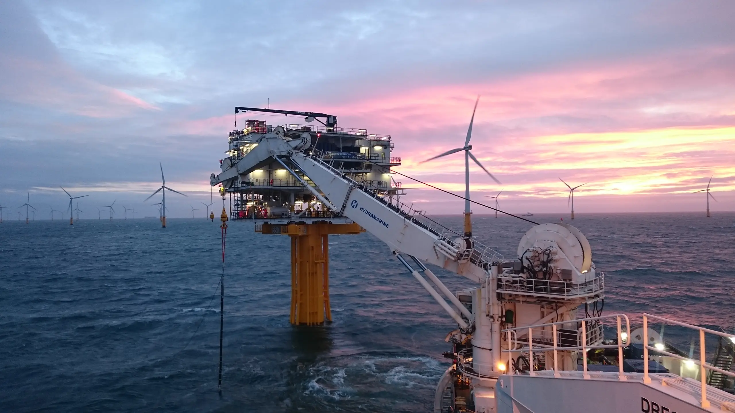 Offshore wind farm with multiple turbines in the ocean at sunset, featuring a large yellow offshore platform and a white crane labeled Hydramarine extending towards it from a vessel.