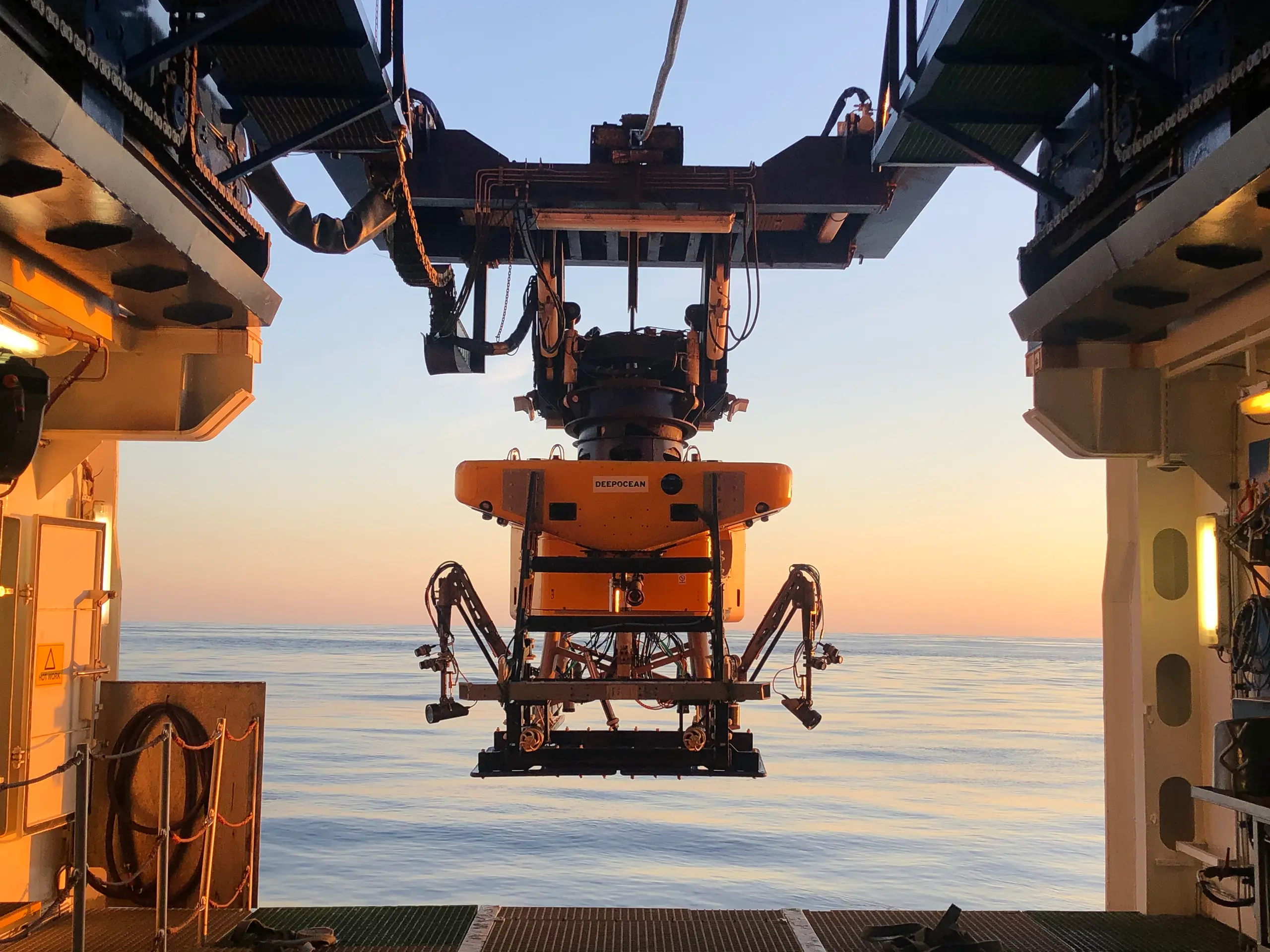 Yellow underwater remotely operated vehicle (ROV) named DEEPOCEAN suspended above the calm sea from a vessel at sunset.