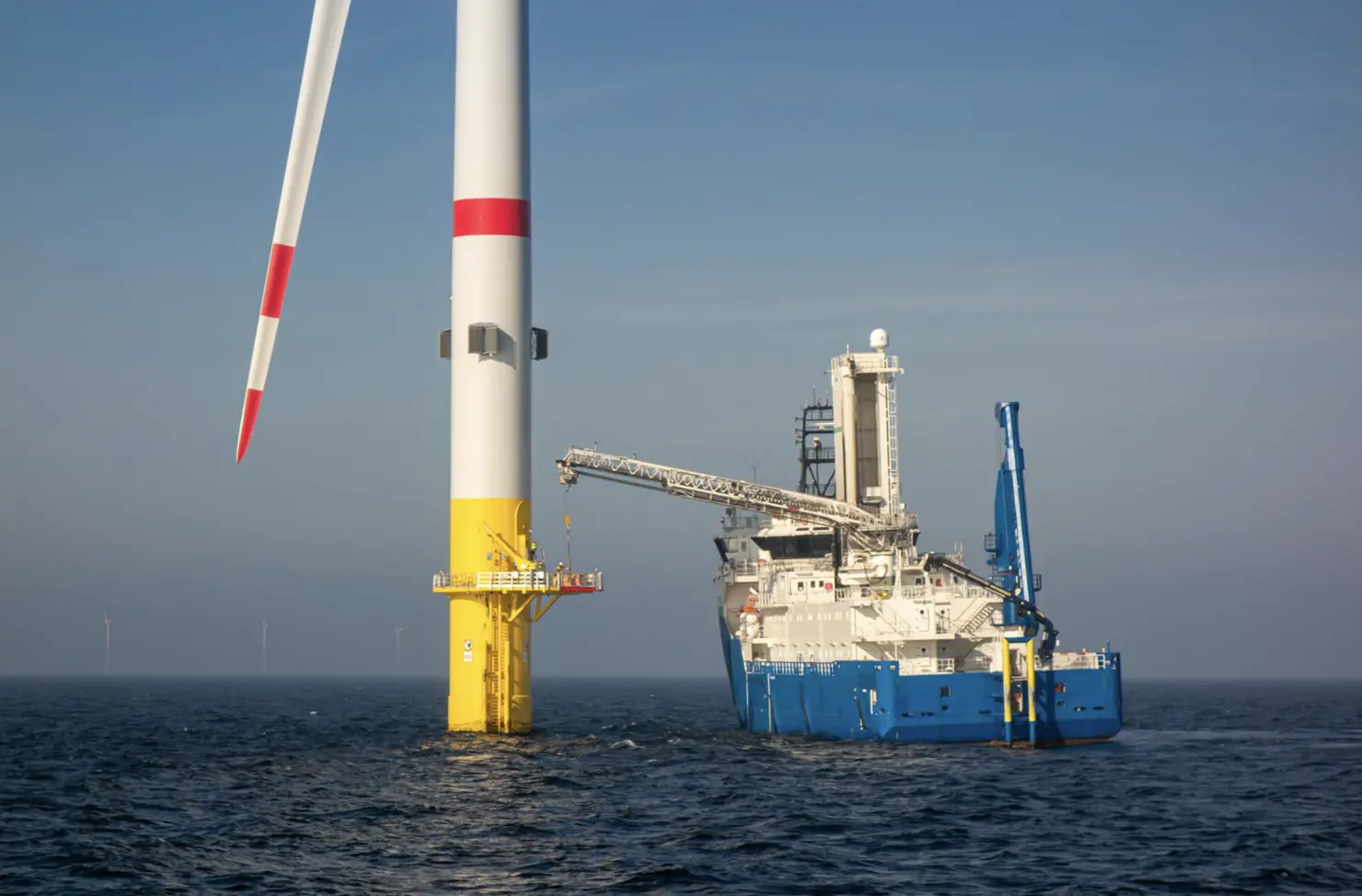 Offshore wind turbine with red and yellow markings on its tower, situated in the sea, next to a large blue and white service vessel equipped with a crane under a clear sky.