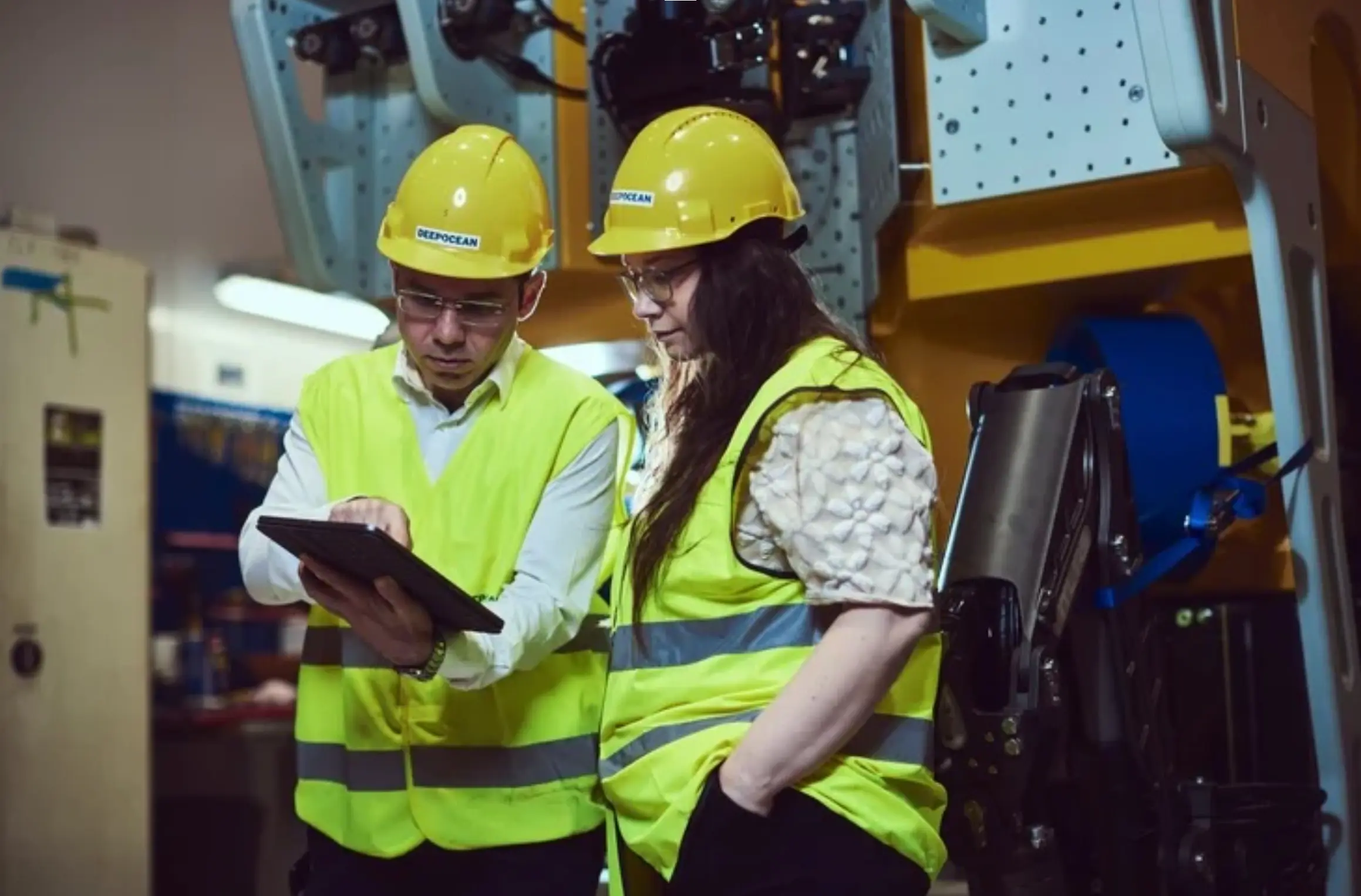 Two industrial workers wearing yellow hard hats and reflective safety vests review information on a digital tablet in a factory setting with machinery in the background.