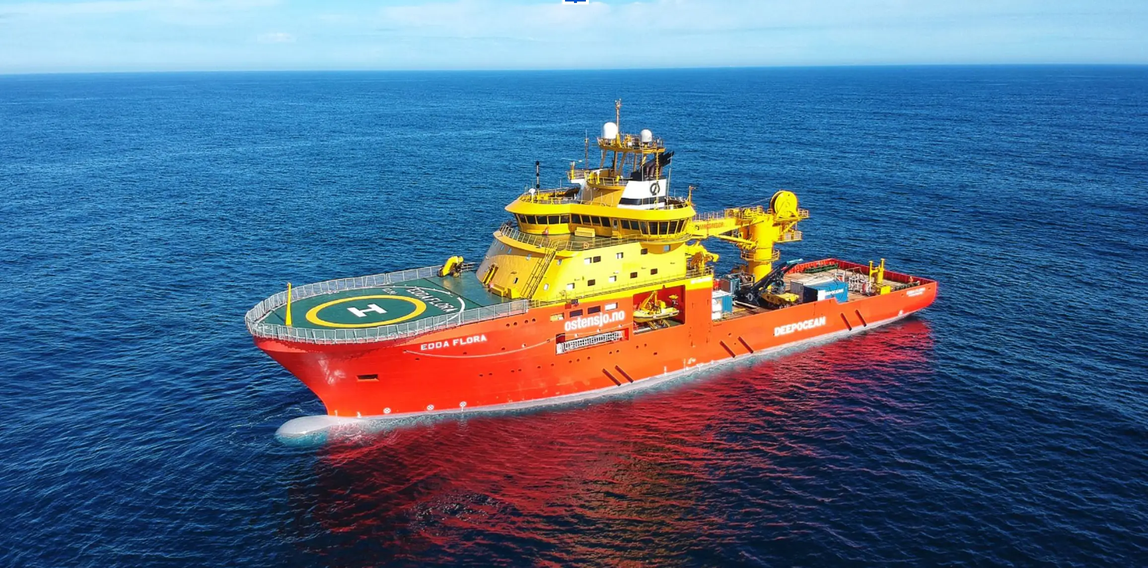 Large red and yellow offshore support vessel named Edda Flora with a helipad, floating on calm blue ocean waters under a clear sky.