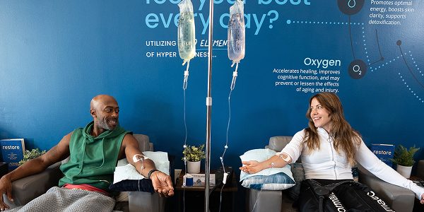 Two clients at Restore Hyper Wellness receiving IV Drip Therapy while sitting in chairs and talking.
