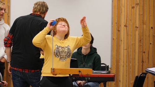 En flicka i kort, rödaktigt hår och gul collegetröja spelar theremin. Hon har armarna uppåt och huvudet lätt bakåtlutat. Hon ler.