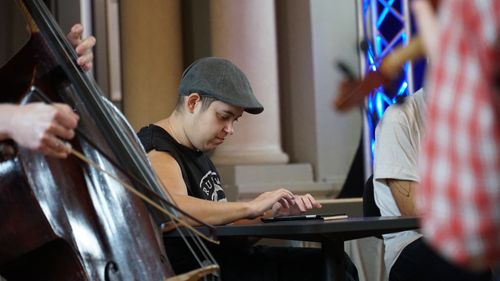 Эвелина из Elefantöra исполняет музыку на планшете, сидя за столом. Вокруг нее видны размытые силуэты других музыкальных инструментов и музыкантов.