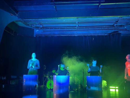 I ett mörkt rum med blått scenljus står tre personer vid vad som ser ut som oljefat. Det är rök omkring dem som är belyst med grönt scenljus.