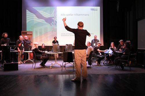 En person syns i förgrunden. I Bakgrunden sitter det andra människor och spelar olika instrument. Mannen i förgrunden håller upp armarna som att han dirigerar.