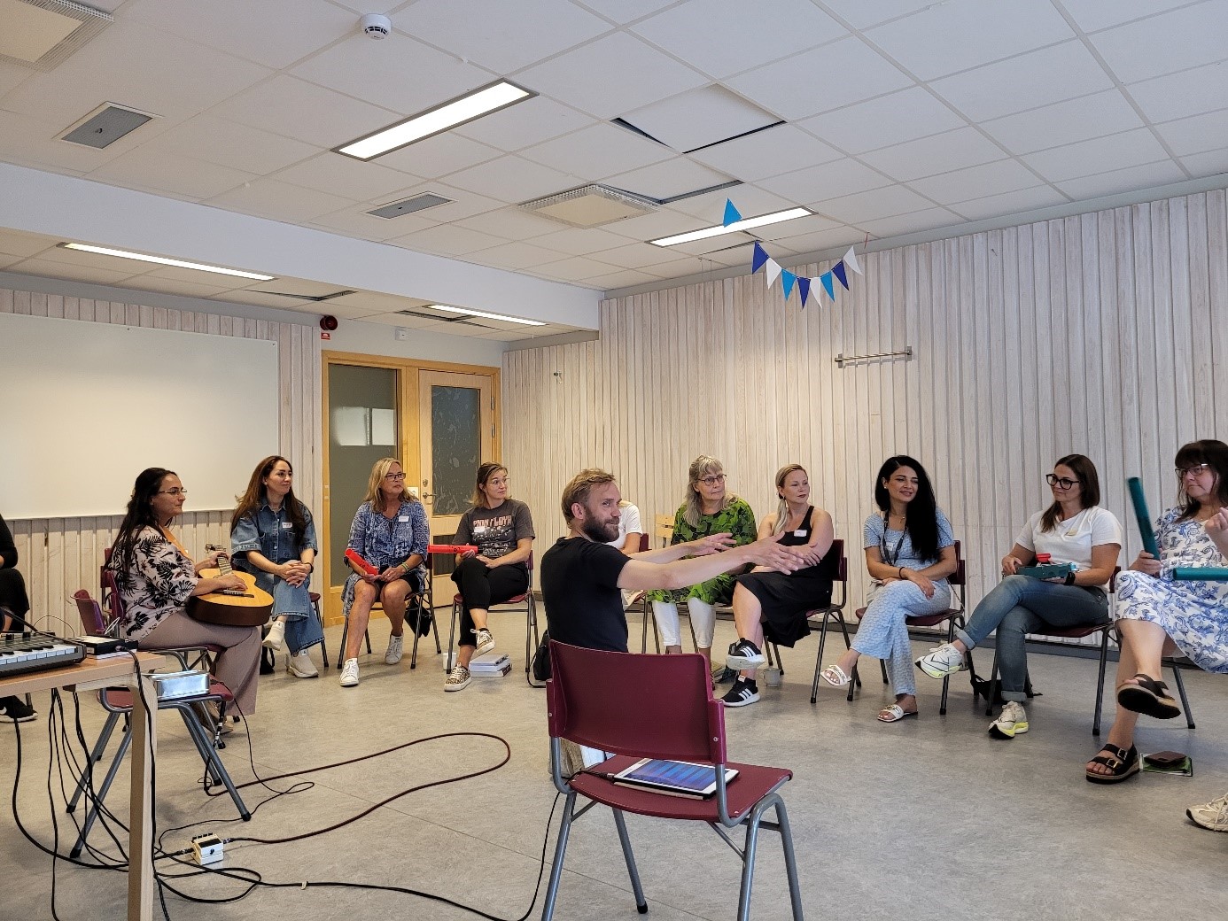 En grupp personer deltar i en musikworkshop i ett klassrum. En person i rullstol spelar ett elektroniskt instrument, medan andra sitter i en halvcirkel med instrument och datorer