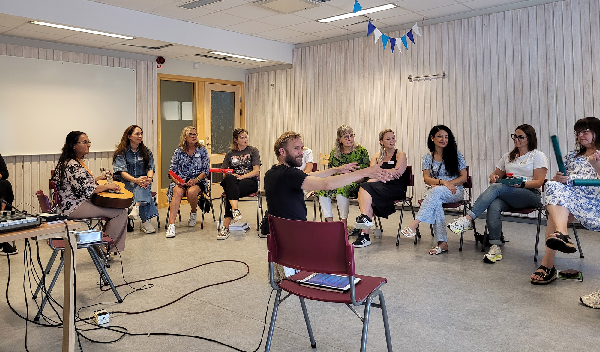 En grupp personer deltar i en musikworkshop i ett klassrum. En person i rullstol spelar ett elektroniskt instrument, medan andra sitter i en halvcirkel med instrument och datorer