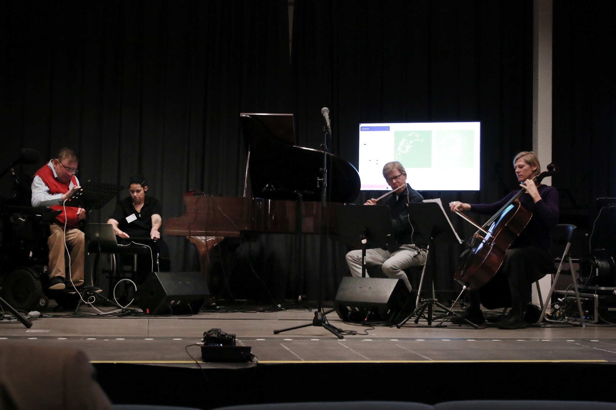 Fyra musiker på scen under MuseIT-sluteventet, inklusive pianospel, cello, flöjt och elektronisk utrustning för distanssamskapande musik. En skärm i bakgrunden visar visuella data som en del av den interaktiva musikupplevelsen.
