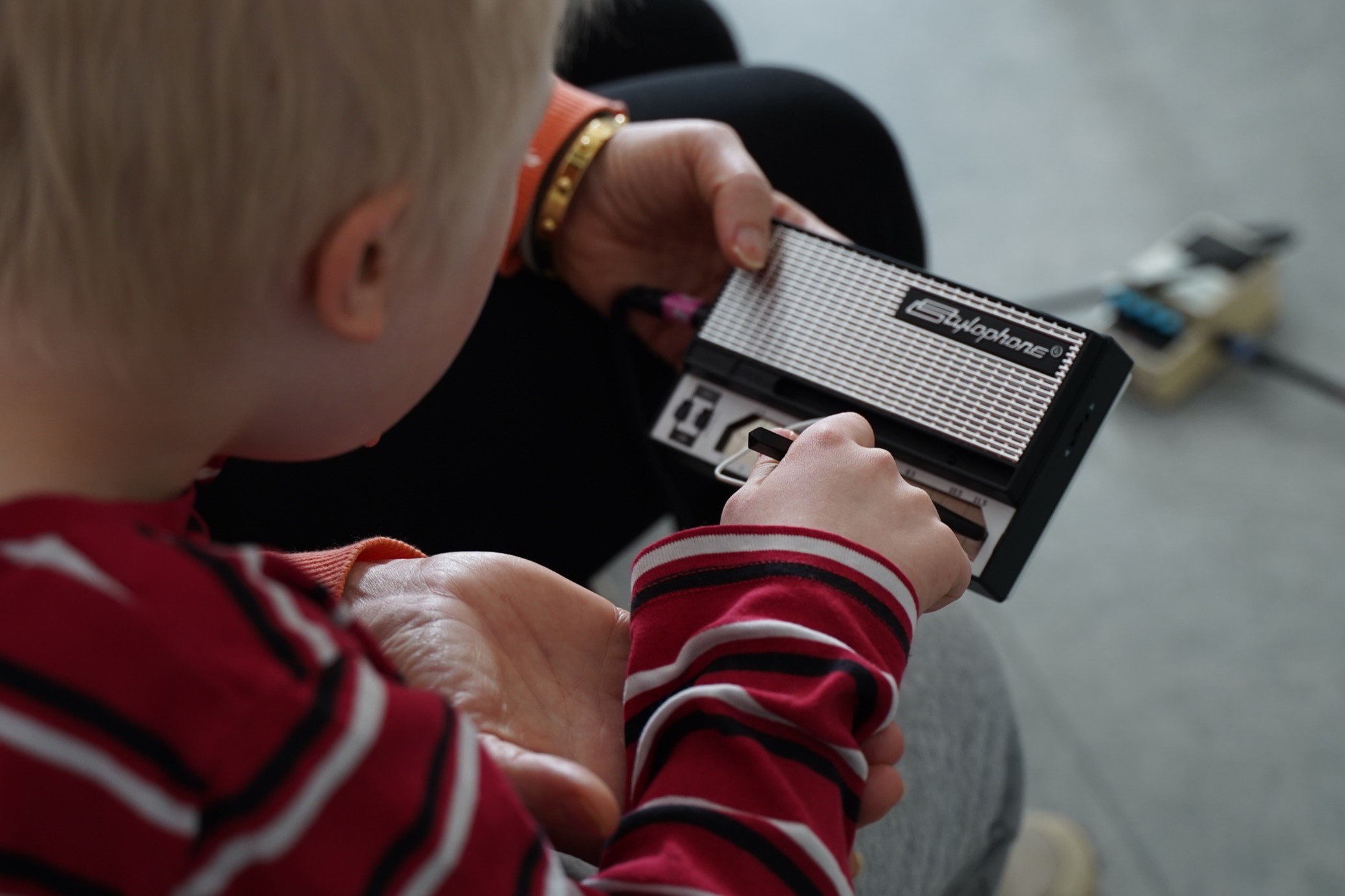 Närbild på ett barn som sitter med en så kallad stylophone i händerna och spelar med en pinne som liknar en penna. Man kan skymta en vuxen bredvid barnet som hjälper att hålla i instrumentet. 