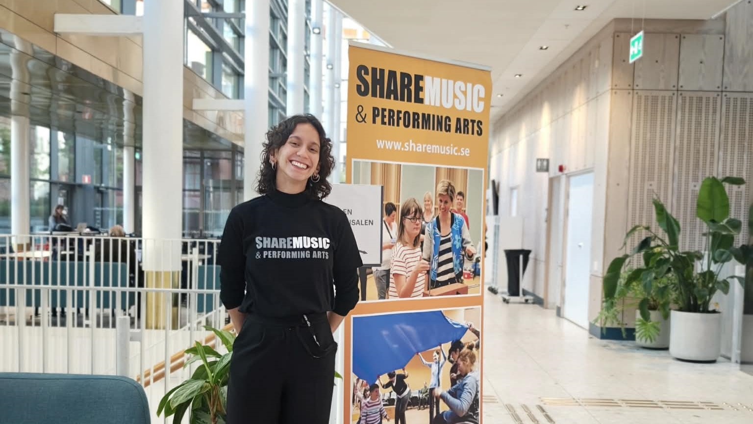 A smiling person wearing a black “ShareMusic & Performing Arts” top stands in a bright indoor venue next to a branded display banner showing photos of inclusive music activities.