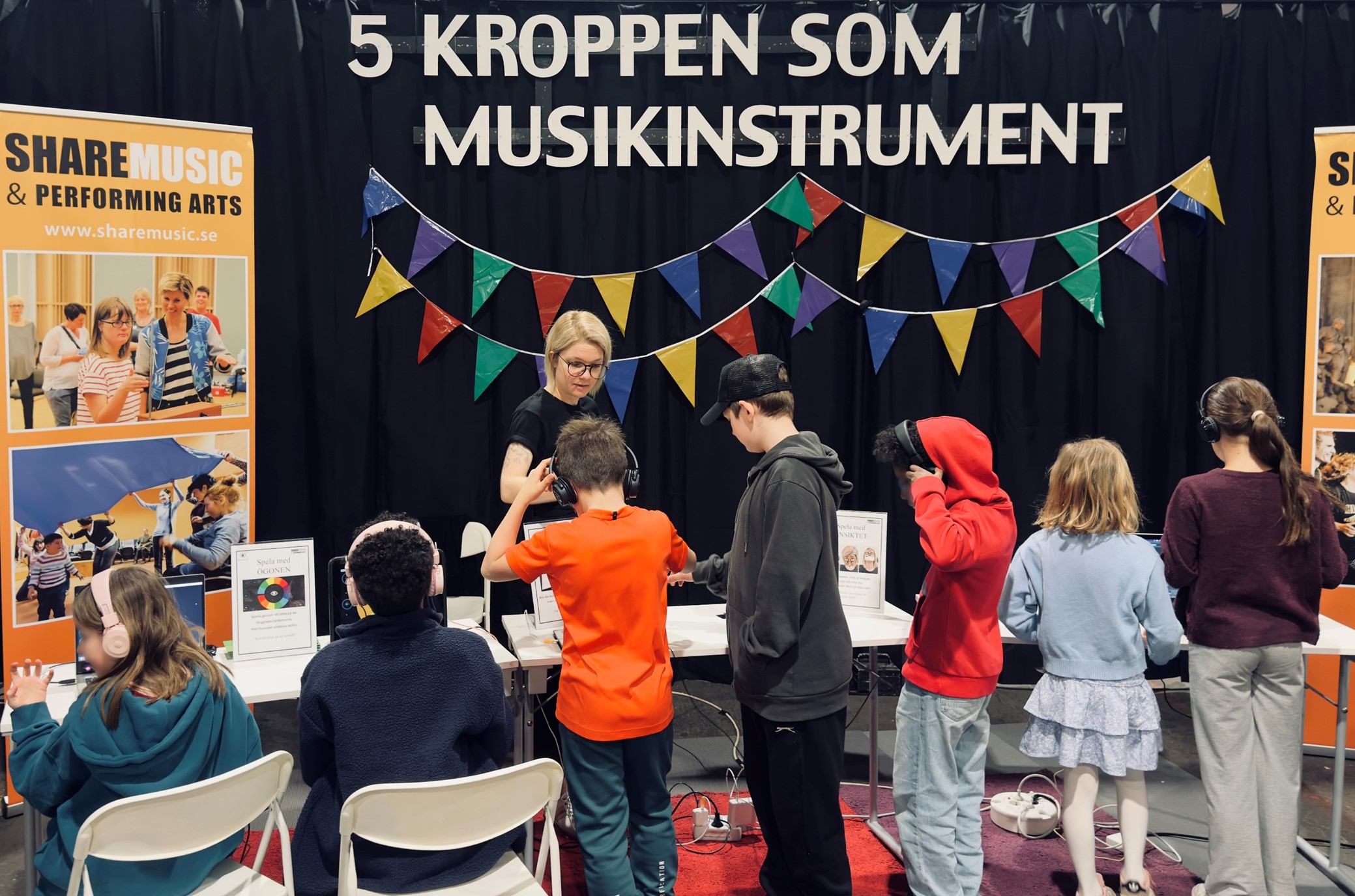 Barn står vid ShareMusic & Performing Arts station på Experimentverkstaden och testar att skapa musik med digitala instrument, medan en ledare visar hur det fungerar.